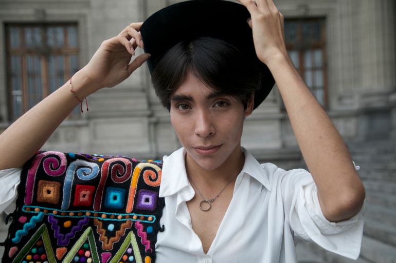 A young Peruvian man lifts his hands to hit hat as he poses for a photo. He wears a crisp white shirt, and over his shoulder is an Indigenous belt, colorfully embroidered.