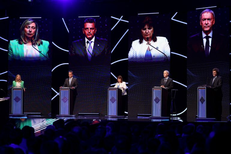 On a darkened stage, five candidates stand on a stage behind podiums, their faces projected onto giant screens behind them.