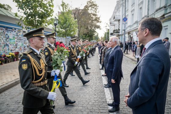 EU foreign ministers line up in front of the Memory Wall of Fallen Defenders of Ukraine in Kyiv. There is a line of soldiers in ceremonial dress holding flowers in front of them. Ukrainian Foreign Minister Dmytro Kuleba is at the front of the photo