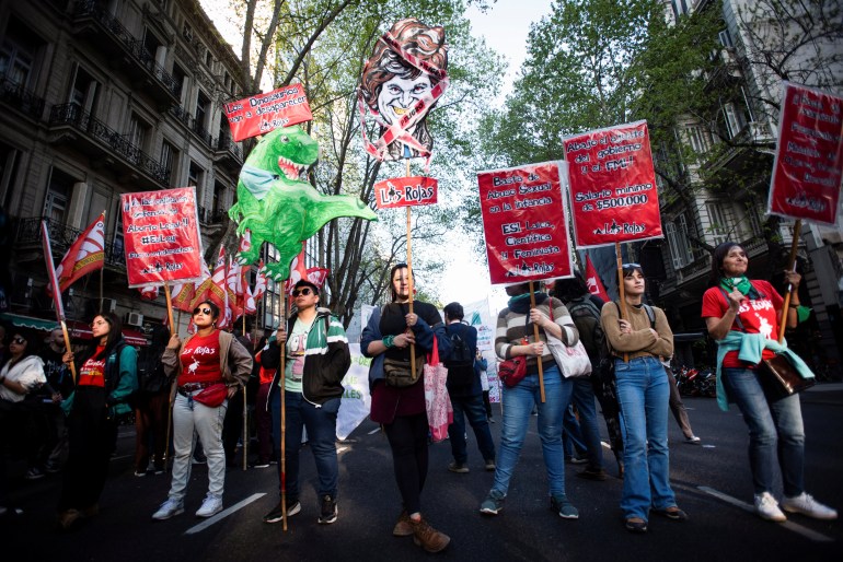 A crowd of demonstrators are on a leafy street in Buenos Aires. Some carry red signs. Another holds a green dinosaur with the message, "The dinosaurs will disappear." And in the middle, a woman holds up a caricature of Javier Milei.