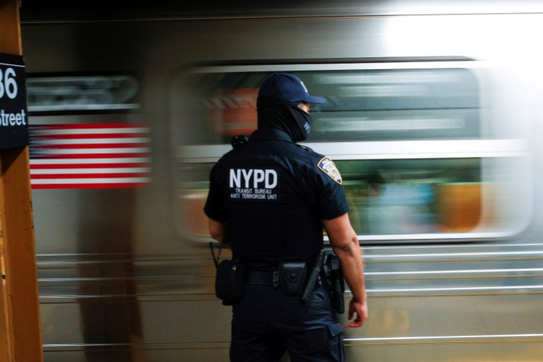 A police officer with "NYPD" written on the back of his uniform stands on a subway platform as a car rushes by.