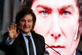 Javier Milei speaks into a microphone and waves. Behind him, a screen displays a giant picture of his face.
