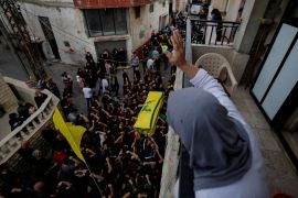 A woman waves from a balcony, as she looks down at the crowd in the street below, who carry a flag-draped coffin.
