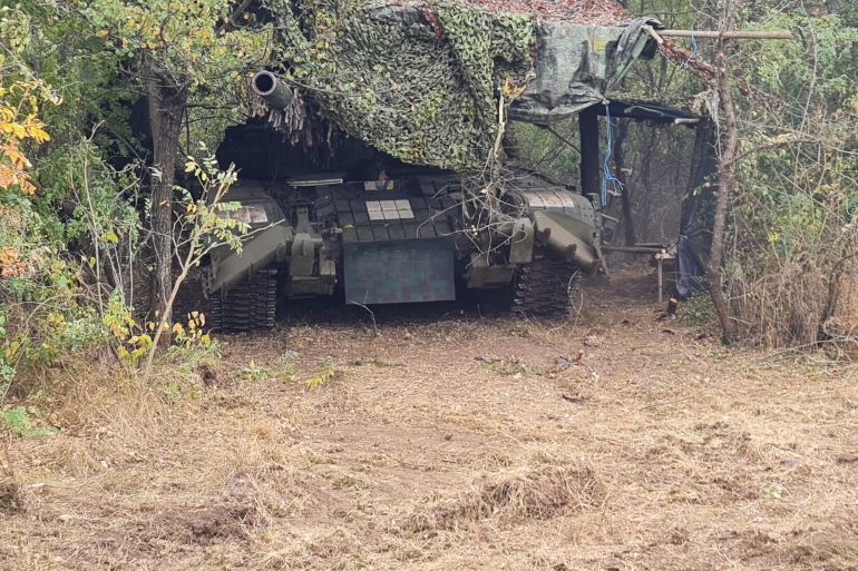 A photo of a tank in between bushes with a blanket to camouflage it on top.
