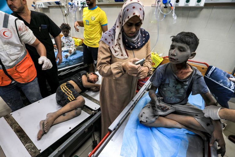 A woman uses a phone while waiting with two children injured in Israeli bombardment