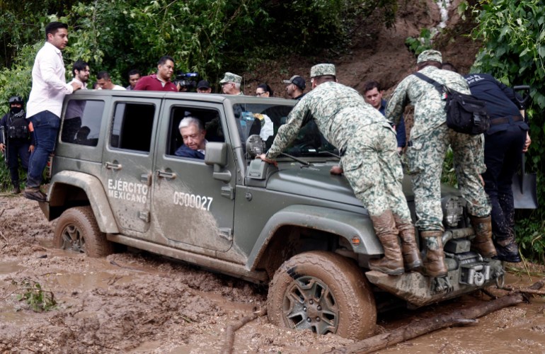 Mexican President Andres Manuel Lopez Obrador in a jeep stuck in the mud. Soldiers are standing on the bonnet. Other people are behind it.