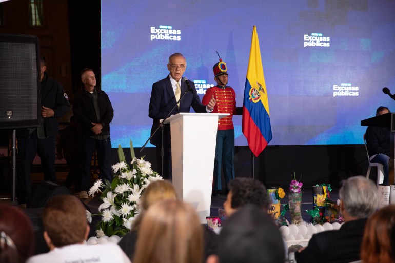 A government official, in a suit and tie, stands behind a podium. Behind him is a soldier in dress uniform, a Colombian flag and a screen.