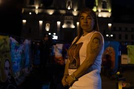 A woman in a white T-shirt stands in a darkened room, where the Colombian government is holding a public event. On her upper arm is a tattoo of a face, representing her 19-year-old son