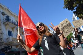 A woman with sunglasses and a black T-shirt flashes a peace sign as she carries a red flag through the streets of Tunis.