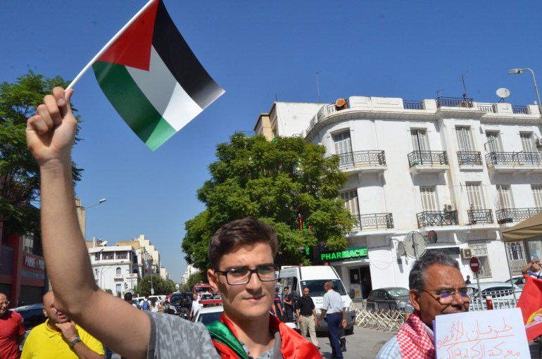 A young male student with glasses waves a Palestinian flag above his head as he marches against a clear blue sky.