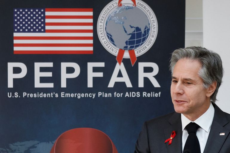 US Secretary of State Antony Blinken sits in front of a large blue banner for PEPFAR, with the acronym and the programme's logo emblazoned on it, alongside an American flag.