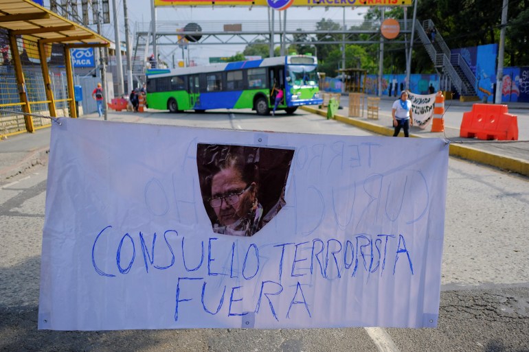 A handwritten banner shows a picture of Guatemala's attorney general and reads "Consuelo terrorista fuera." In the background, a bus can be seen blocking the road.