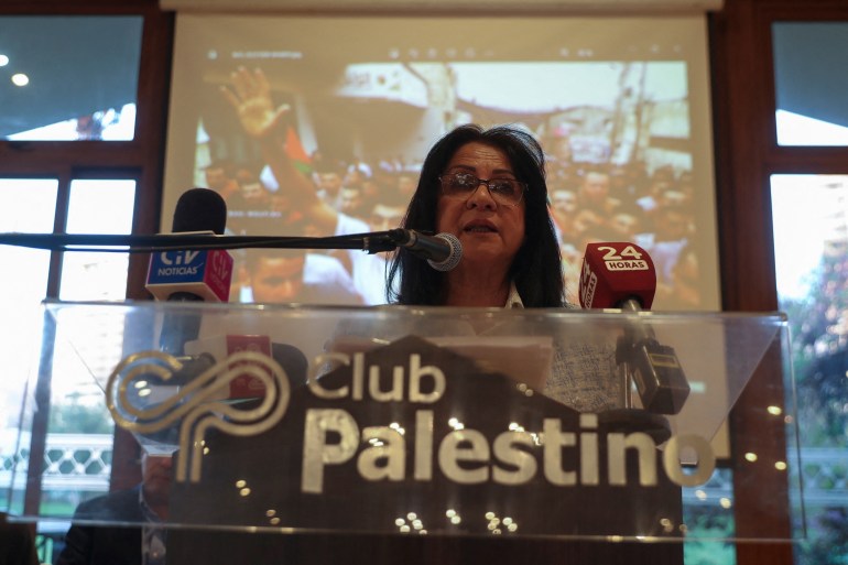 A woman stands at a glass podium engraved with the name "Club Palestino," as she speaks into a microphone.