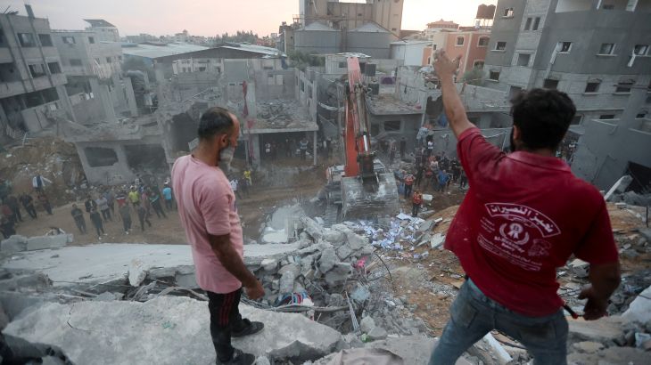 Palestinians searching for survivors in the rubble of a collapsed building. Two men are standing on the debris. An excavator is below. There are more damaged building behind