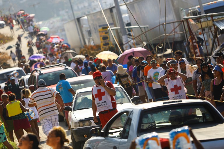 An Acapulco street is crowded with cars and a long line of people, stretching toward the horizon, as aid workers — visible with a white apron emblazoned with a red cross — stand watching.