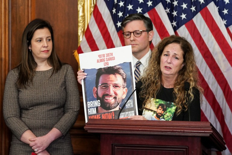 Doris Liber speaks at a wooden podium in Congress, holding up a poster-sized photo of her missing child. Mike Johnson and Else Stefanik stand behind her, near a set of American flags.