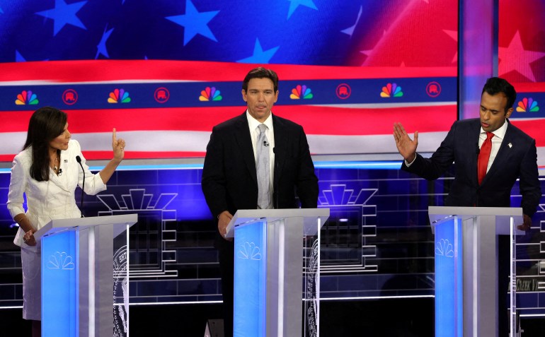 Behind three glass podiums, three Republican candidates stand: Nikki Haley in a white suit set, Ron DeSantis in a dark suit and light-coloured tie and Vivek Ramaswamy in a dark suit and red tie. Ramaswamy and Haley speak and gesture, while DeSantis licks his lips.
