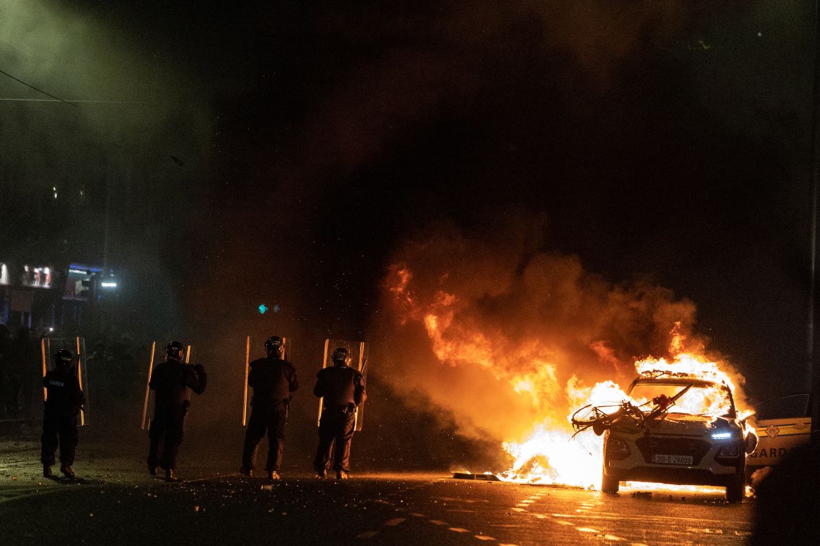 Riot police officers face down demonstrators next to a burning police car