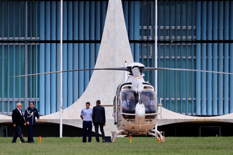 Brazilian President Lula da Silva walks alongside a military official, as a service member in a blue shirt and another person wait to escort him onto a helicopter. The swooping architecture of the Alvorada palace in Brasilia is visible behind them.