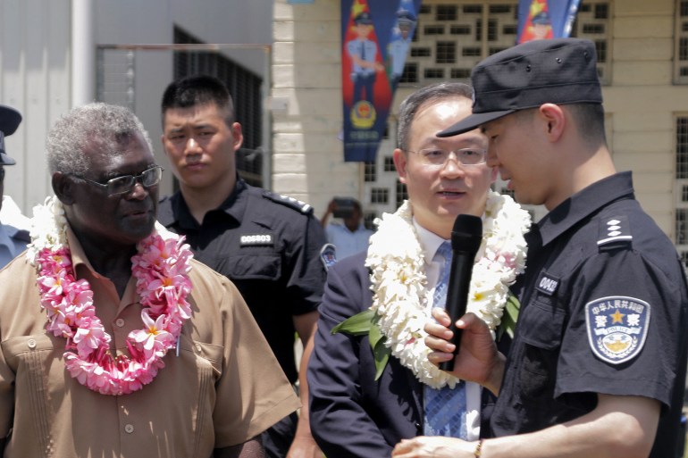 Solomon Islands Prime Minister Manasseh Sogavare (L) looks on with Li Ming (2nd R), China's ambassador to the Solomon Islands listening to a speech by a police officer. They have garlands around their necks.