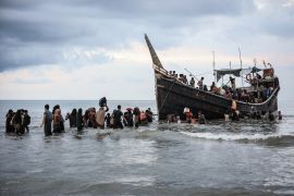 Refugees walking back through the water to their boat after being refused to land. The boat has a prominent bow.