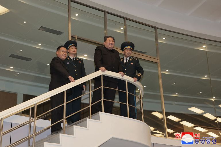 Kim Jong Un standing on a balcony at a Pyongyang aerospace centre He is accompanied by military officers. He is smiling.
