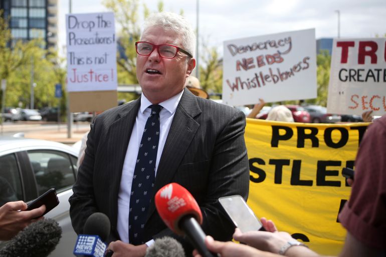 A man in a suit with several journalist microphones in front of him stands in front of signs about whistleblowers