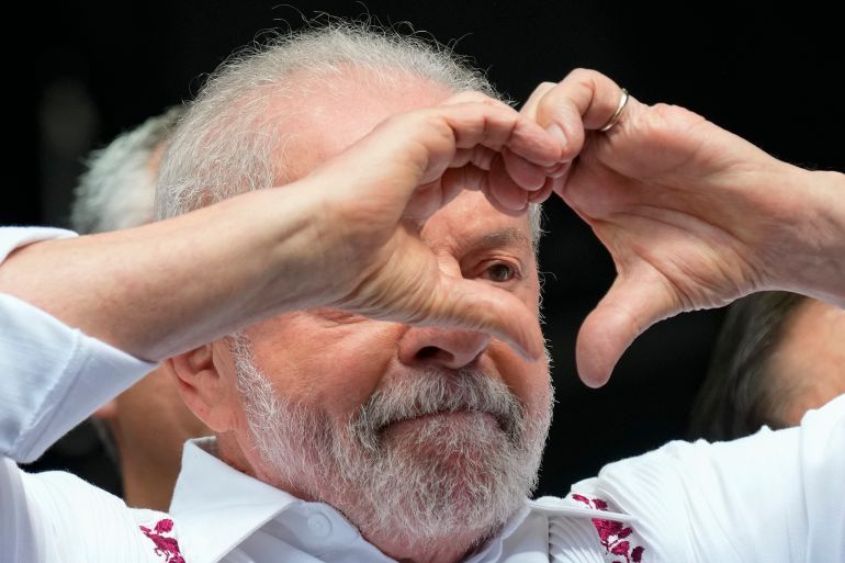 Brazilian President Lula da Silva raises his two hands to form the shape of a heart. He wears a white collared shirt in this close-up photo.