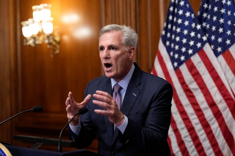 Kevin McCarthy gestures with both hands outstretched in a wood-paneled room decorated with US flags.