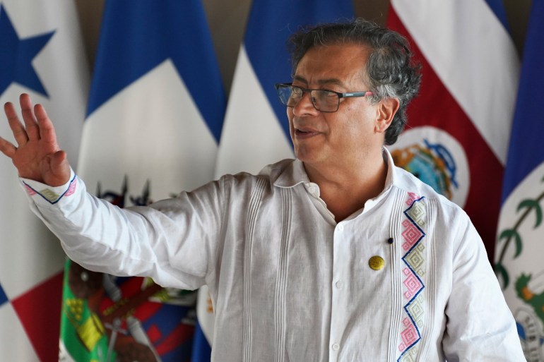 Dressed in a white embroidered shirt, President Gustavo Petro raises an arm in gesture as he speaks before a row of flags.