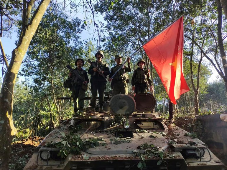 Soldiers from the Myanmar National Democratic Alliance Army standing on top of a capture Myanmar military tank. They have hung their flag on the tank. They are surrounded by trees.
