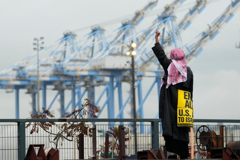 A protester, wearing a keffiyeh on their head and holding a protest sign, holds a fist in the air. In the background are dock cranes indicative of a port.