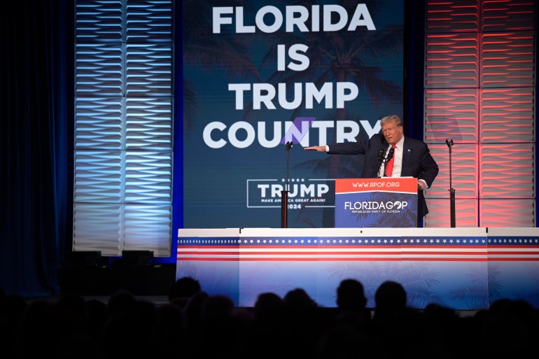 Donald Trump, in a dark suit and red tie, points from behind a podium at a campaign rally. Behind him is a sign that reads: Florida is Trump country.