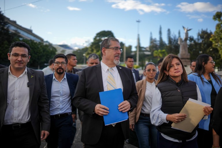 Guatemala Bernardo Arevalo wears a suit and holds a blue folder as he walks outside with his vice president Karin Herrera, who wears a puffer vest and long-sleeved shirt.