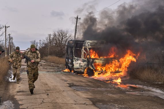 A burning bus on the side of the road as two Ukrainian soldiers walk past.