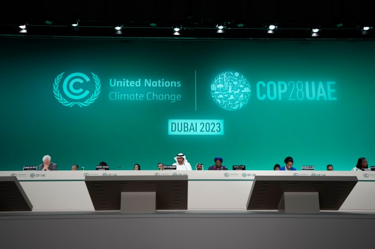 A panel table sits along the length of a stage in Dubai, behind which officials sit. The background of the stage is a green screen with logos and slogans like "Dubai 2023," "United Nations Climate Change" and "COP28 UAE."
