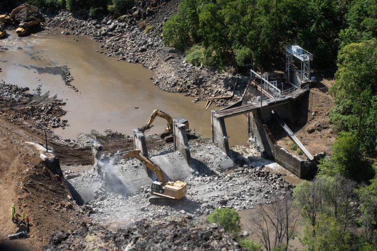 Heavy-duty excavators tear down the concrete walls of a dam on the Klamath River, seen from above amid a hilly landscape covered in trees and brush.