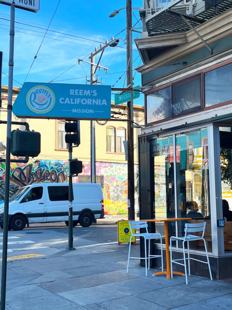 The exterior of Reem's, a Palestinian-owned restaurant in San Francisco. A white van passes on the road nearby, as diners can be seen inside a restaurant window, under a sign that reads: "Reem's California, Mission"