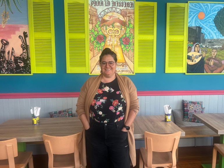 Zaynah Hindi of the restaurant Reem's stands in front of wooden tables and a painted wall in her Mission District location.