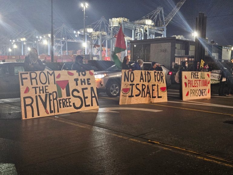 Protesters form a line across a rain-slick road leading to a port. They hold up three large banners. "From the river to the sea," one reads. Another reads, "No aid for Israel."