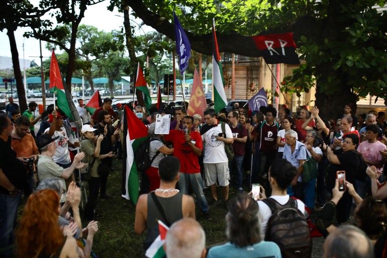 Protesters gather outside the US consulate in Rio de Janeiro. One speaks into a microphone and lifts up a sign. Others hold up Palestinian flags.