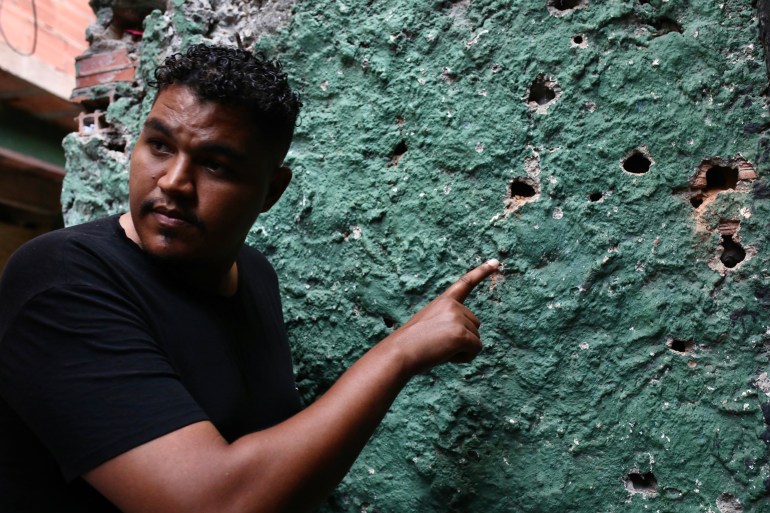 Cosme Felippsen, wearing a black T-shirt, points to a wall pockmarked with bullet holes.