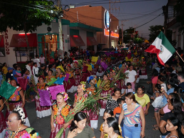 Muxes — members of Mexico's third-gender community — carry bouquets and wear traditional dresses during a night parade where bystanders wave Mexican flags. 