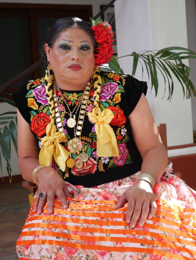 A muxe sits in a wooden chair, wearing an orange ribbon skirt and an elaborately stitched black top, decorated with flowers. The muxe's hair is braided with yellow ribbons and topped with beautiful red flowers.