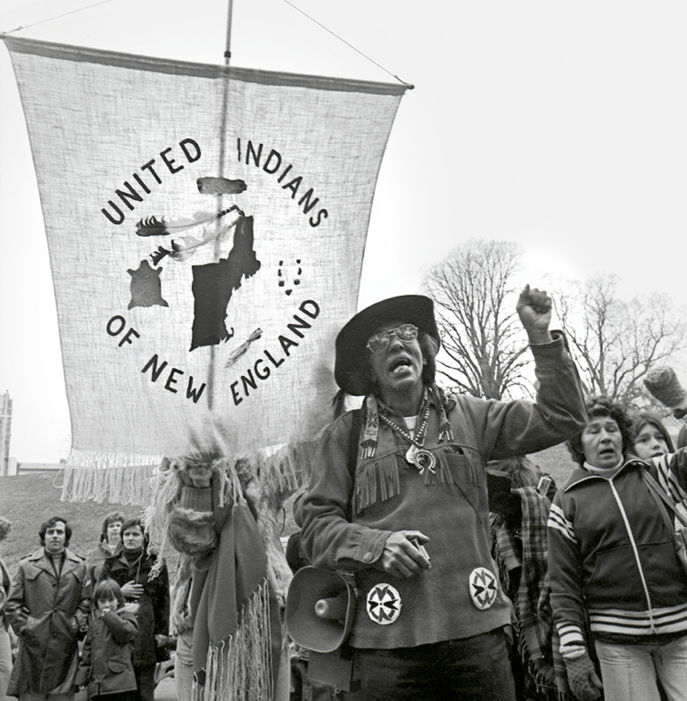 Wamsutta Frank James lifts a fist in solidarity as he marches in a black-and-white 1972 photo. A banner behind him reads: United Indians of New England.