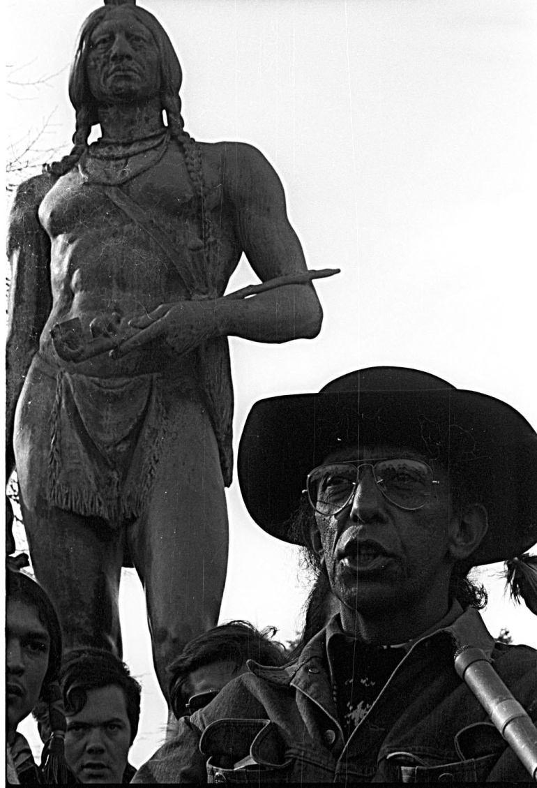 Wamsutta Frank James, wearing glasses and a broad hat, speaks in front of the statue of Wampanoag leader Massasoit in a black-and-white photo