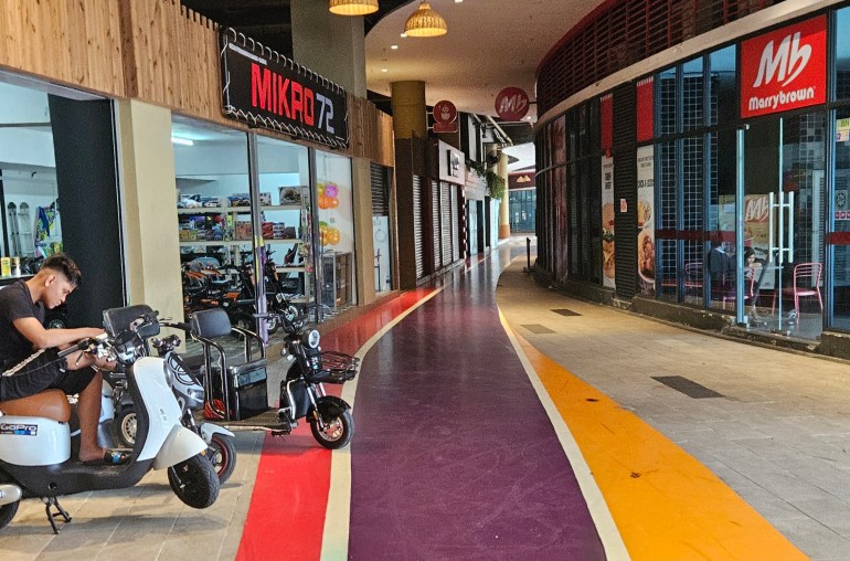 A man sitting on a scooter outside a shop that offers the bikes to rent. There is.a fast food outlet opposite but the area is deserted.
