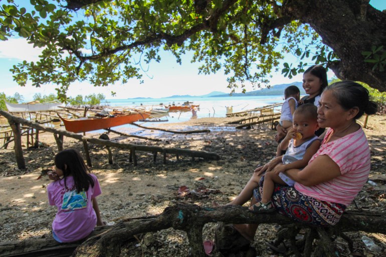 Jecel Espina-Pedel sitting with her family by the sea. She is standing and holding a baby. The others are seated on a log.