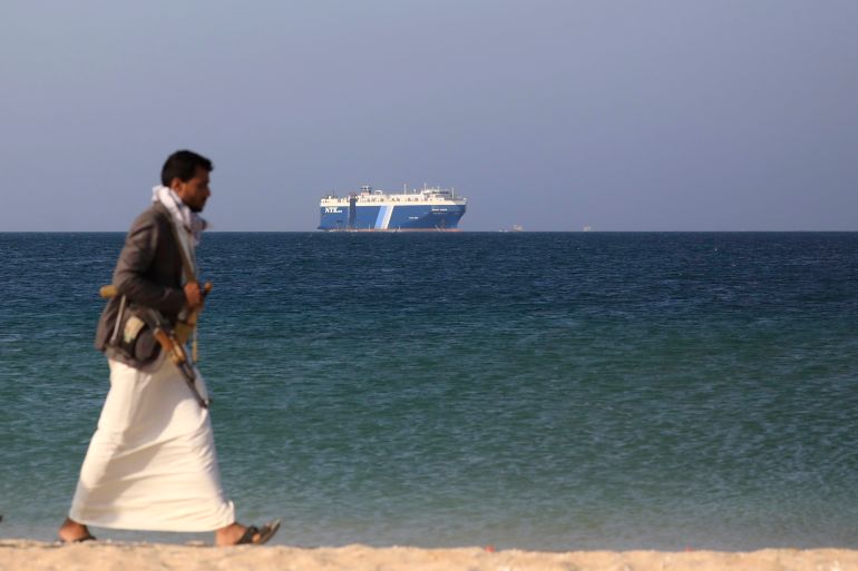 An armed Houthi fighter walks through the beach with the Galaxy Leader cargo ship in the background, seized by the Houthis offshore of the Al-Salif port on the Red Sea in the province of Hodeidah, Yemen