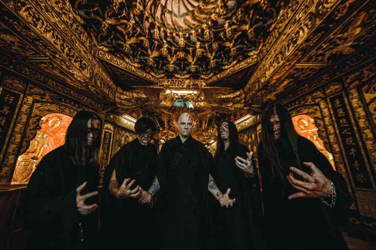 Dharma's 2021 line-up, from left to right: Andy Lin (guitar), Bull Tsai (bass), Joe Henley (vocals), Jon Chang (ex-guitar), Jack Tung (drums). They look like they are in a room in a temple. They are wearing black robes and scowling. 